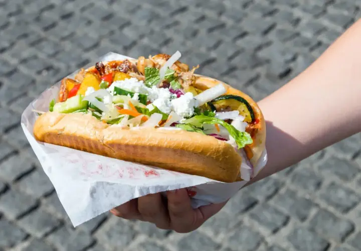 Ręka trzymająca dużą bułkę z kebabem, pełną mięsa, warzyw i sera feta.