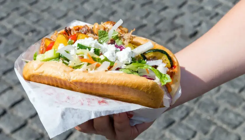 Ręka trzymająca dużą bułkę z kebabem, pełną mięsa, warzyw i sera feta.